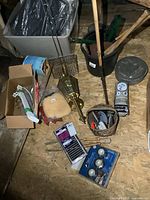 Wide view of lot items arranged on plywood floor including brass coach light, campfire cooking basket, metal brackets, valve, wooden bats, floor register, tongs, and boxed lighting items.
