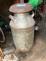 Front-side view of vintage metal milk can with lid, showing embossed writing and aged rust.