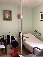 Full view of the white adjustable mobility assist pole positioned between a hospital-style bed and a wall, showing grab bar and padding.