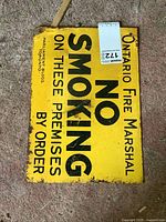 Full view of yellow metal no smoking sign with black text reading Ontario Fire Marshal NO SMOKING ON THESE PREMISES BY ORDER. Visible wear and rust around edges.