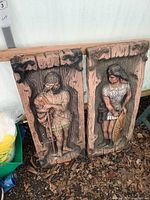 Pair of resin wall hangings showing Viking warrior and shield maiden figures with detailed carved and painted features and rustic wood background.