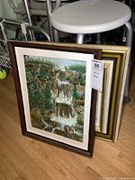 Two framed oil on canvas paintings leaning against a stool and other items, showing the larger waterfall painting in front and the smaller painting partially visible behind.