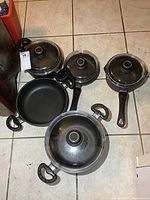 Overview of pots and pans arranged on tile floor, showing metal cookware with black handles and glass lids.