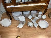 Wide shot showing all dishes grouped on floor near cabinet, includes plates stacked, bowls, cups, creamers, sugar bowls, platters, and serving bowl