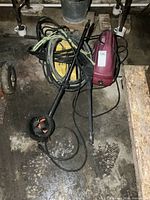 Two red Kodiak 3100 electric pressure washers with hoses and attached spray wands, one with circular brush attachment visible, placed on a basement floor.