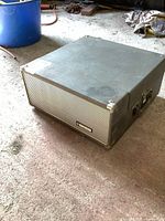 Closed view of vintage portable record player with textured exterior and reinforced corners, placed on a floor.