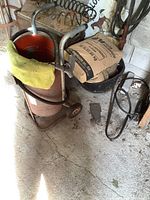 Front angled view of gravity fed sandblaster showing red barrel with orange interior, black hose wrapped around, mounted on metal wheeled cart with handles. Several bags of abrasive sand nearby.