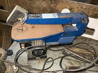 Side view showing full scroll saw with blue body, motor housing, and table surface. Power cord visible.