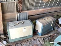 Two vintage sewing machines inside damaged hard cases placed on wooden surface in garage area. The cases show dirt, wear, and damage. Machines untested.