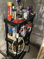 Black resin shelving unit with various spray paints, household liquids, and miscellaneous items on three tiers in basement setting.