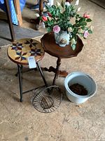 Photo showing vintage side table with scalloped edges holding porcelain planter with artificial flowers, folding stool with painted star pattern, small white porcelain planter, and wire basket arranged on floor.
