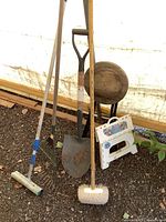 Lot of garden tools including a shovel, rake, squeegee, brush, plastic step stool, and wooden folding stool against a tent background.