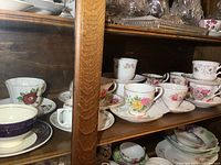 Photo showing multiple teacups and saucers with floral and blue/gold patterns inside a wooden cabinet shelf.