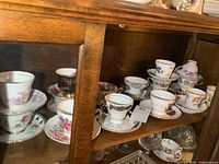 Photo showing 10 teacups and saucers arranged on a wooden shelf, various floral and patterned designs visible