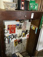 Wide shot of pegboard with various small tools and hardware hanging, including C clamps, hooks, marrettes, and pipe fittings in plastic bins above.