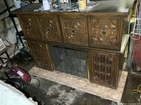 Front view of vintage bar cabinet showing carved wood doors, electric fireplace grill, and marble base with wear.