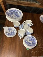 Photo of 9 cups and saucers along with stacks of plates showing blue countryside pattern on white china.