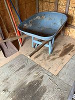 Side angled view of a vintage metal wheelbarrow with wood handles on a wooden floor inside a shed, showing wear and paint chipping.