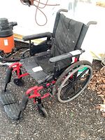 Side view of the Elite manual wheelchair showing red metal frame, black padded seat and backrest, footrests, and wheels. Some dust and dirt visible on plastic and fabric surfaces.