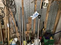Wide view of assorted yard tools including shovels, rakes, roof rake poles, sprayer, bucket with parts, and souvenir plate in a shed.
