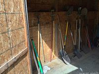 Full view of various yard tools including shovels, hoes, rakes, garden fork, and sledge hammer leaning against shed wall