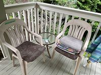 Two beige resin patio chairs, round glass side table, cushions, partial view of striped umbrella