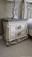 Nightstand showing ornate carved front, two drawers with decorative handles, and marble top with Greek key pattern