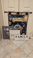Front view of all four decorative signs and wall art arranged on the floor leaning against a white door. Includes a large space-themed tapestry and three rectangular wood signs with Home, Adventure, and Family themes.