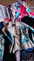 Top view showing mixed women's tops in various colors including beige floral cardigan, black and peach tops, colorful fleece jacket with pink and blue panels, striped shirts.