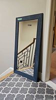 Photo showing full view of the modern dark blue wood framed mirror standing against a white wall next to staircase and gray patterned rug.