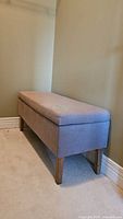Grey fabric upholstered bench ottoman placed against a beige wall and carpeted floor. Shows side perspective.