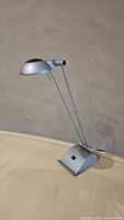 Full view of the silver adjustable desk lamp showing the lamp head, adjustable arms, and base on a light wooden surface.
