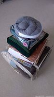 Stack of various pillows and folded bedding items, some in clear protective cases and packaging.