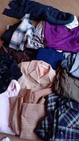 Pile of women's clothing showing various jackets, sweaters, skirts, and tops in different colors and patterns on wooden floor.