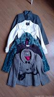Four women's jackets arranged vertically on wooden floor: dark gray, white, teal fuzzy, and grey patterned jacket.