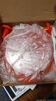 Top view of the chicken shredder device wrapped in plastic inside a cardboard box, showing round orange plastic with shredding teeth and suction pads.