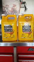 Two yellow 1-gallon jugs of Harris Scorpion Killer indoors on a metallic red surface—visible labels and handles.