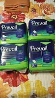 Four unopened packs of Prevail underpads arranged on a floral bedspread, each pack contains 25 units.