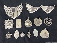 Top view of all 14 pewter pendants laid out on black surface showing diverse shapes and designs including abstract, floral, and nautical.