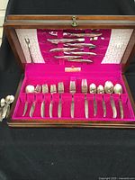 Full view of flatware set in wooden box with pink felt lining showing multiple knives, forks, and spoons arranged in slots.