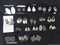 Photo showing full lot of 18 pairs of pewter earrings arranged in rows on black background, varied shapes and styles.
