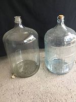 Photo showing both clear glass carboys side by side on a dark background, the larger one without stopper and the smaller with a cork stopper.