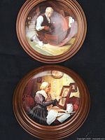 Photo showing two Norman Rockwell collector plates in wooden wall mounts, one with an elderly man working on a wooden project and another with an elderly woman painting a photo.