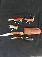 Five cutters and knives including Swiss Army multi-tool, additional multi-tools, two wooden handled lock blade knives with leather sheaths all laid on black fabric