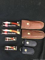 Four lock blade cutters in original boxes with two brown leather sheaths and two smaller black leather sheaths on black background.