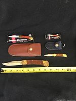 Four lock blade pocket cutters shown with two leather sheaths and two original black boxes labeled 'Stainless Steel Blade'. Measuring tape shows large knives approximately 7.5 inches open with about 4 inch blade. Handles are wood with brass bolsters and metal lock mechanism.