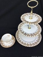 Full view showing three-tier plate stand with gold floral decoration and matching teacup and saucer on black background