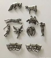 Overhead photo showing 8 distinct pewter brooches including animals, human figures, and a fairy, alongside 2 pewter hair clips with theatrical mask designs, all laid out on a flat light-colored surface.