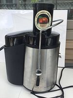 Front and side view of the Big Boss electric juicer showing stainless steel and black plastic construction, extra large feeder with labeled sticker, and metal locking arm.