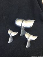 Three whale tail sculptures displayed on a black fabric background, showing their shapes and polished metal surfaces.